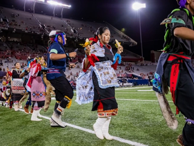 Hopi dancers performing a corn dance in traditional tribal clothing on a football field with stadium lights shining down