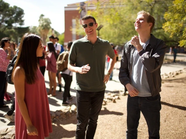 Three University of Arizona students stand and chat in a sunny outdoor area near Old Main. The student on the left wears a sleeveless pink dress, the student in the middle wears sunglasses and a dark green polo shirt, and the student on the right wears a gray T-shirt and jacket. Other students can be seen talking and walking in the background beneath tall trees.
