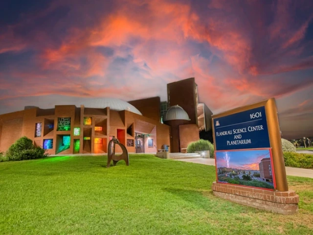 The exterior of the Flandrau Science Center and Planetarium at sunset. The building is constructed with red brick and features modern architectural designs, including a large dome that hints at its planetarium function. A series of rectangular windows are illuminated with various colors, creating a vibrant spectrum on the building's facade.