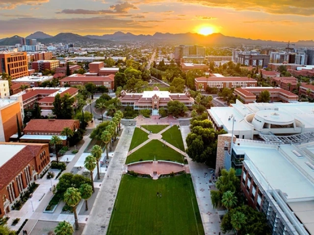 Aerial view of the University of Arizona campus at sunset, with Old Main at the center, the grassy Mall stretching toward the mountains in the distance, and the sky glowing with orange and purple clouds.