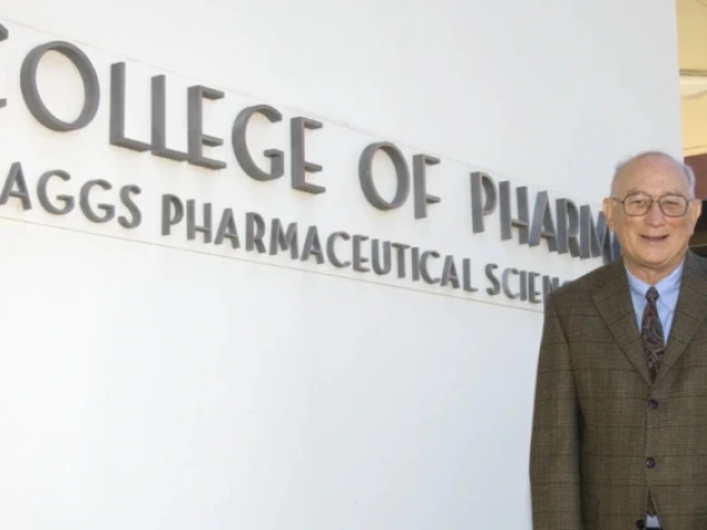 Jack R. Cole, wearing a brown plaid suit and patterned tie, stands smiling in front of a white wall with large lettering that reads "College of Pharmacy Skaggs Pharmaceutical Sciences."