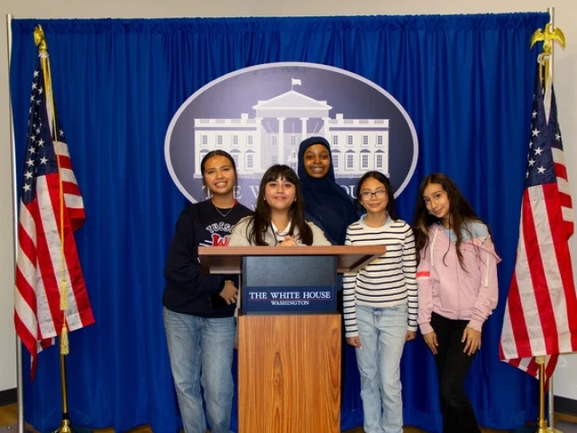 Five young people standing together behind a podium, which indicates "The White House Washington." The group is posed in front of a royal blue curtain displaying a large emblem of the White House. Two American flags are positioned on either side of the curtain, each with a gold-colored eagle finial at the top. The individuals are smiling and casually dressed, with one wearing a hijab.