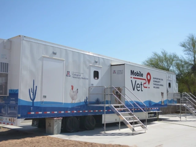 A large mobile veterinary clinic trailer against a clear blue sky. The trailer is white with blue accents and features images of animals, including a chicken and a dog. The left side displays the logo and text for the University of Arizona College of Veterinary Medicine. Metal stairs with railings lead to entrances on the side of the trailer. Various graphics, including cacti, are painted along the bottom.