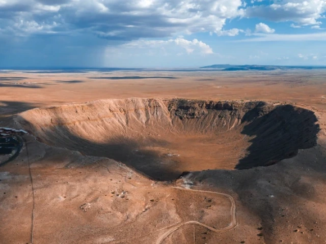 an aerial view of a meteor crater under cloudy blue skies on a vast desert landscape